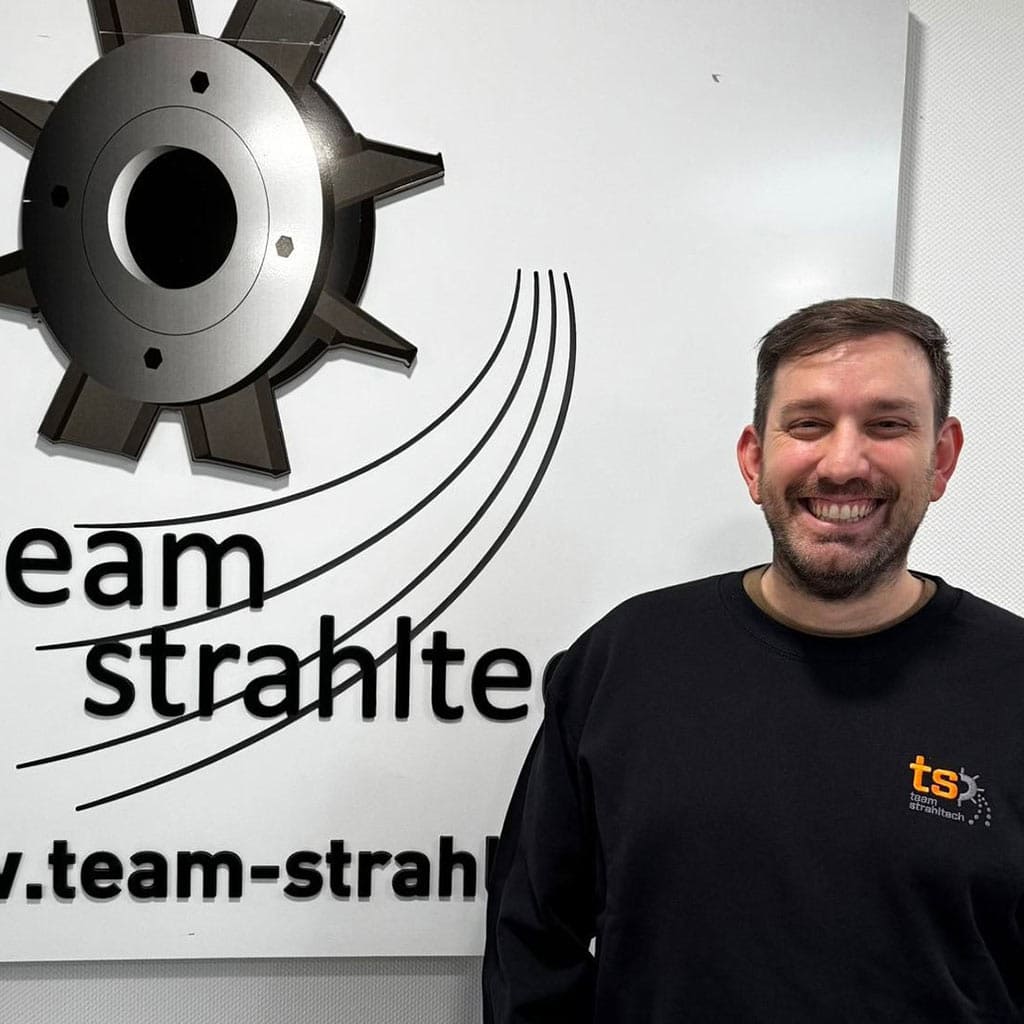 Smiling man with dark hair, wearing a black sweatshirt, stands by a sign with gear-like design.